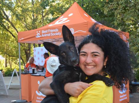 Smiling person holding the black puppy she adopted in front of a Best Friends booth