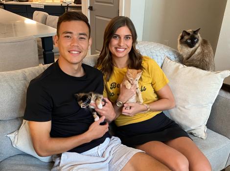 Sam Junqua and Emma Besier sitting on a couch holding foster kitten with a cat beside them on the armrest