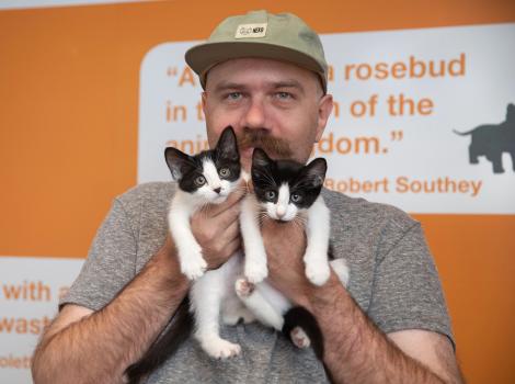 Foster Tyler Lisonbee wearing a hat and gray T-shirt, holding two black and white kittens in front of him