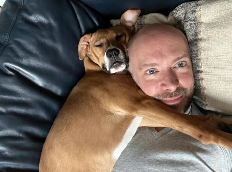 Volunteer David Sprague lying in a bed with a brown and white dog sleeping and snuggled next to him