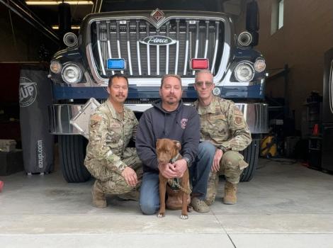 Ruthie the dog with three people (two in military uniforms) in front of a large vehicle