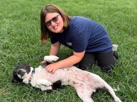 Person kneeling on the grass next to a dog who is lying on her back